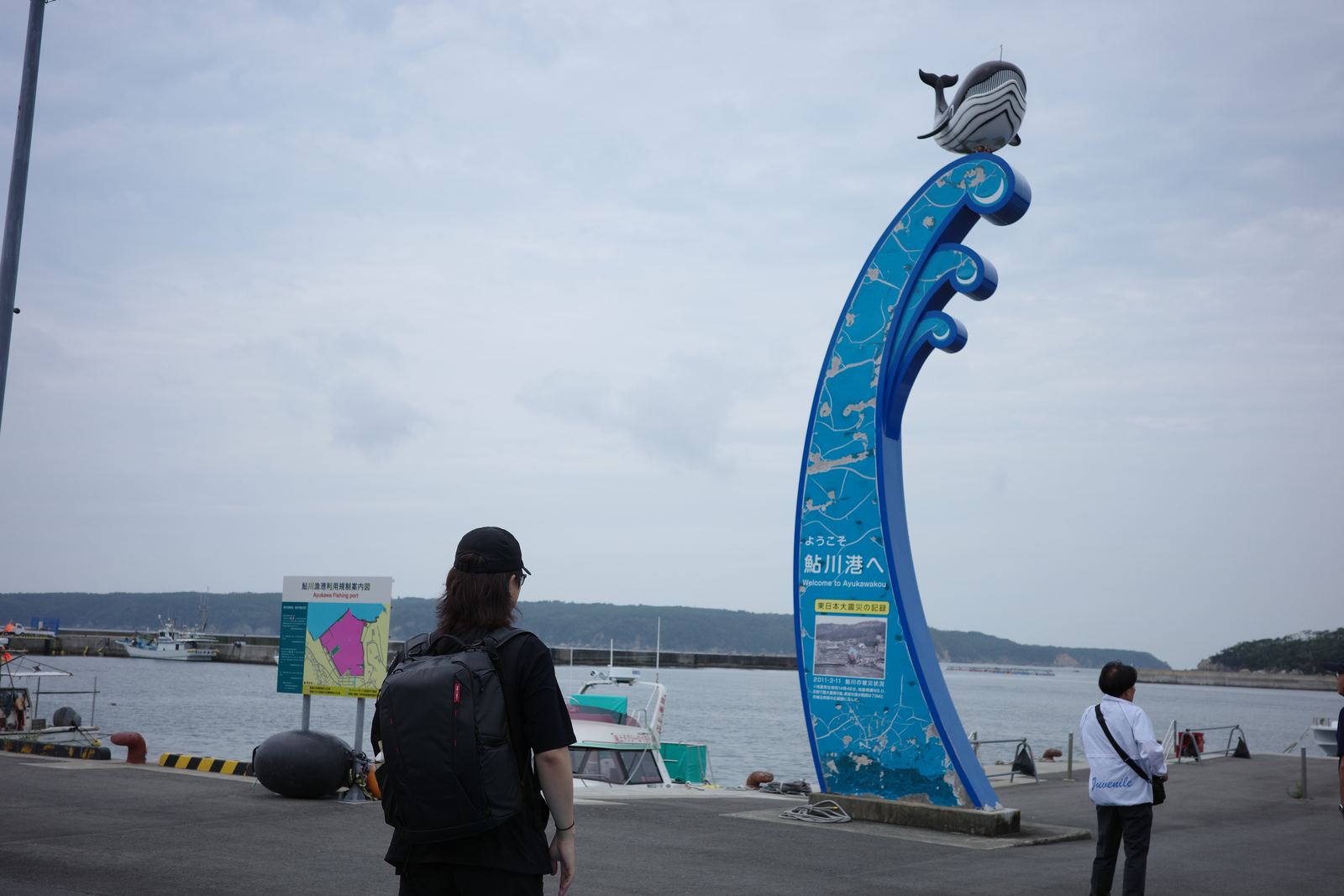 クジラのオブジェが載った青い波形のモニュメント「ようこそ鮎川港へ」。手前にリュックを背負った人物の後ろ姿