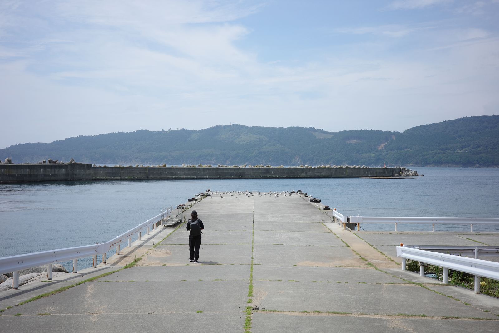 晴れた空の下、金華山港の桟橋を歩く人物。奥に防波堤と対岸の山並み
