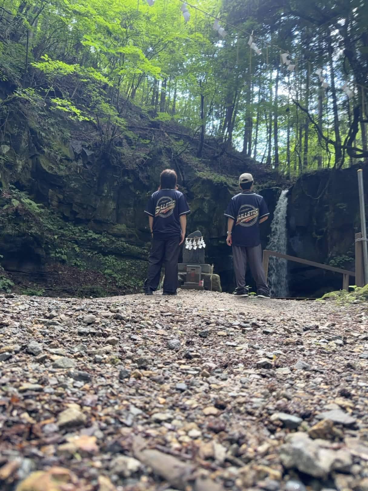 ネイビーのベースボールシャツを着たBULLETISの2人が、小僧不動の滝と石碑の前に後ろ姿で立っている。地面すれすれの低いアングル