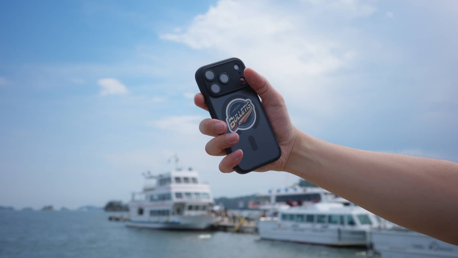 松島湾の遊覧船を背に掲げたスマートフォン。背面にBULLETISのサブロゴのステッカーが貼ってある
