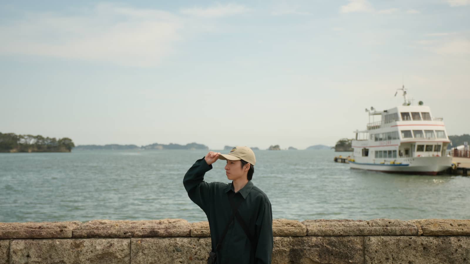 キャップをかぶったTANAYANが、遊覧船の浮かぶ松島湾を眺めている