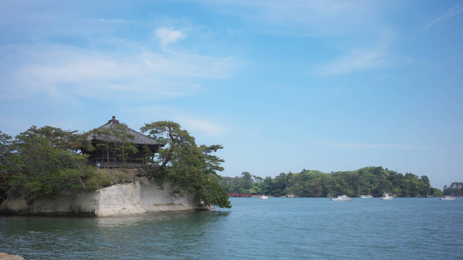 青空の下、松島湾に浮かぶ島。岩の上に建つお堂と、奥に遊覧船が見える